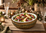 A woven gold and brown bowl filled with a colorful salad, set on a burlap placemat on a wooden table with holiday decorations, glassware, and a candle in the background.