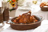 A roasted whole chicken in a clear glass baking dish with a brown woven basket-style base, set on a white tablecloth in a dining setting with cutlery, bread, and flowers in the background.