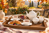 A styled autumn outdoor tea time setup on a woven tray, featuring a white ceramic teapot, sugar bowl, creamer, two cups of tea, a croissant, and a bowl of berries, with a blurred garden background.
