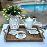 Tea set with teapot, cups, and saucers on a woven tray with a vase of flowers in the background.