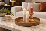A round woven tray holding three lit candles in a styled living room setting with a marble coffee table, sofa, and decorative pillows.